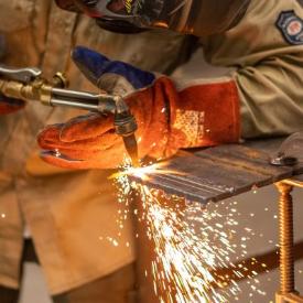 Welding student wearing safety gear and demonstrating how to use a tig welder, sending sparks across the picture.